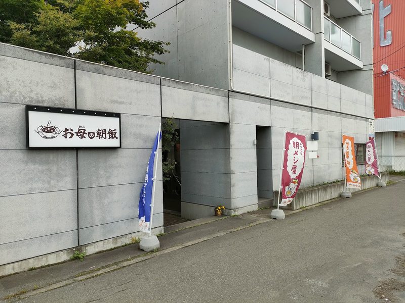 コンクリートの建物に掲げた「お母の朝飯」の看板とのぼり